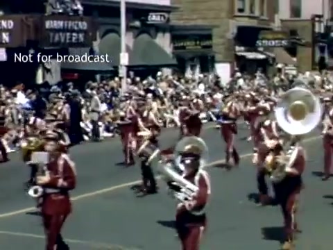 The still captures a lively scene from the Holland, Michigan Tulip Festival Parade in the late 1940s. A marching band, dressed in uniforms featuring red and white colors, is performing in the street, with musicians playing brass instruments. A large audience of spectators, seated along the sidewalks, enjoys the parade amidst historical buildings in the background. The vibrant atmosphere reflects the festive spirit of the event.