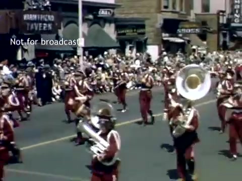 The still captures a lively scene from the Holland, Michigan Tulip Festival Parade in the late 1940s. A marching band is prominently featured, with musicians dressed in red uniforms, playing various instruments including a tuba. In the background, a crowd watches and local buildings are visible, enhancing the festive atmosphere of the parade. The overall mood is celebratory and community-focused.