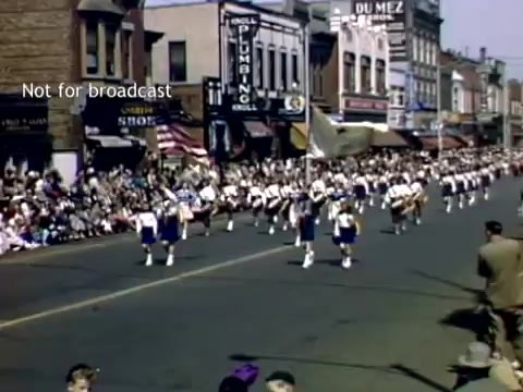 The still captures a vibrant scene from the late 1940s Holland, Michigan Tulip Festival Parade. The image shows a marching band in uniforms marching down a street lined with spectators. Historical storefronts are visible in the background, adorned with signage. The atmosphere is festive, reflecting the community spirit of the parade.