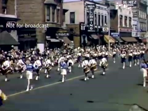 The still captures a vibrant scene from the late 1940s Holland, Michigan Tulip Festival Parade. A marching band is shown parading down a city street lined with spectators. The musicians, dressed in coordinated outfits, play instruments while onlookers, including families, enjoy the festivities. The backdrop features a mix of storefronts, reflecting the architectural style of the time. The colorful scene emphasizes the celebratory atmosphere of the festival.