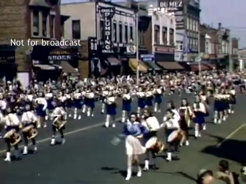 The still captures a vibrant scene from the late 1940s Holland, Michigan Tulip Festival Parade. A lively procession of marchers in blue and white outfits, likely a marching band, fills the street. The backdrop features a range of storefronts, showcasing the architecture of the era. Crowds line the street, enjoying the festive atmosphere. The image is colorful, reflecting the celebratory nature of the event.