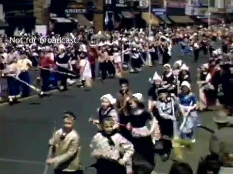 The still captures a lively scene from the late 1940s Holland, Michigan Tulip Festival Parade. It features a colorful procession with participants in traditional attire, including floral dresses and wooden shoes. Families and children are joyfully participating, with a bustling crowd in the background enjoying the festivities. The street is lined with shops, contributing to the vibrant atmosphere of the parade.