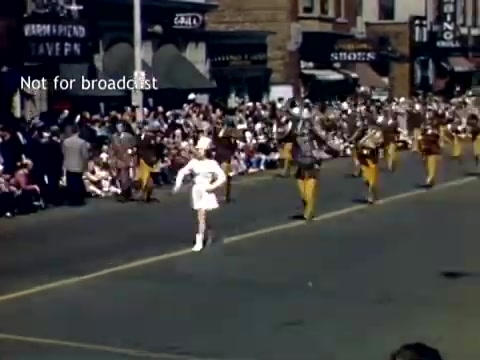 The still captures a vibrant scene from the Holland, Michigan Tulip Festival Parade in the late 1940s. A line of marching musicians, dressed in uniforms, performs along a street lined with spectators. A figure in a white outfit takes the lead, likely a majorette or leader of the marching band. The atmosphere is festive, with onlookers enjoying the parade. Historic buildings and storefronts frame the background, enhancing the quaint, community-focused vibe of the event.