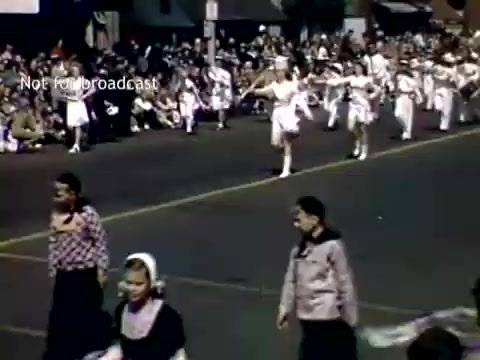 The still depicts a vibrant scene from the late 1940s Holland, Michigan Tulip Festival Parade. You can see a procession featuring a group of performers, likely dancers, in white costumes. They are parading along a street lined with spectators who are seated on the curb, enjoying the festivities. The atmosphere appears festive and community-oriented, capturing a moment of celebration and cultural heritage.