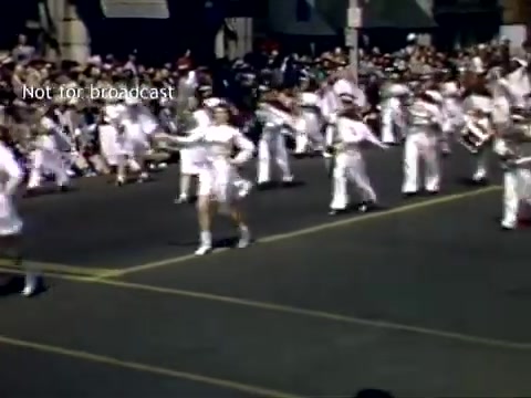 The still depicts the Holland, Michigan Tulip Festival Parade from the late 1940s. It showcases a lively procession featuring participants dressed in white, marching down a street lined with onlookers. The atmosphere is festive, capturing the community spirit of the event.