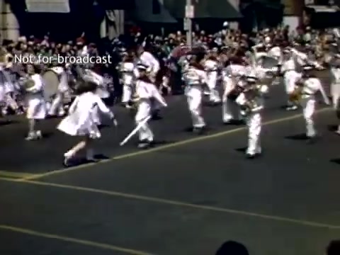 The still depicts a lively scene from the late 1940s Holland, Michigan Tulip Festival Parade. It features a group of performers, likely a marching band or drill team, dressed in white uniforms. They’re parading down a busy street lined with spectators, some holding umbrellas. The festive atmosphere is enhanced by the colorful clothing and the energy of the participants as they perform in the parade.