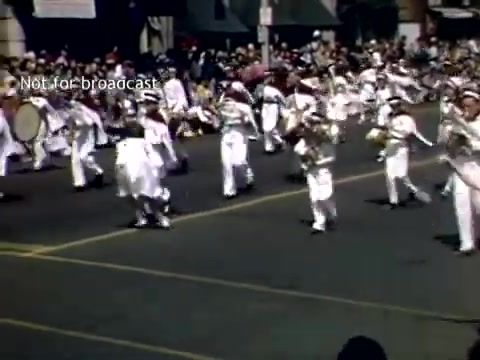 The still depicts a lively scene from the Holland, Michigan Tulip Festival Parade in the late 1940s. A marching band is performing, with members dressed in white uniforms, creating a vibrant and festive atmosphere. The street is lined with onlookers, reflecting the community's engagement with the celebration. The overall tone is joyful, capturing the spirit of the parade during that time period.