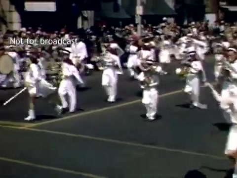 The still captures a lively scene from the Holland, Michigan Tulip Festival Parade in the late 1940s. A marching band, dressed in white uniforms, plays instruments as they parade down a street filled with spectators. The vibrant atmosphere is enhanced by bright colors and a sense of community celebration.