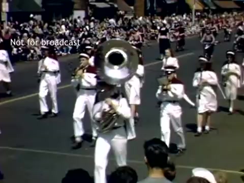 The still captures a lively scene from the late 1940s Holland, Michigan Tulip Festival Parade. A marching band is prominently featured, with musicians dressed in white uniforms and playing various instruments, including a tuba. The street is lined with spectators, eagerly watching the procession. Colorful decorations and festive elements likely enhance the atmosphere of the celebration.
