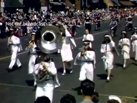 The still captures a vibrant scene from the Holland, Michigan Tulip Festival Parade in the late 1940s. A marching band is prominent, featuring musicians in white uniforms, including a tuba player at the forefront. Spectators line the street, eagerly watching the procession, while colorful storefronts and festive elements of the parade enhance the celebratory atmosphere. The image reflects the spirit of community and festivity characteristic of the annual event.