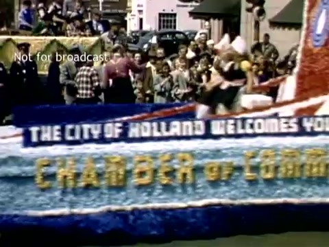 The still captures a vibrant scene from the late 1940s Holland, Michigan Tulip Festival Parade. A colorful float, adorned with floral decorations, prominently displays the message welcoming attendees from the 'Chamber of Commerce.' A crowd of spectators, dressed in period attire, gathers along the street, enjoying the festive atmosphere. The setting reflects a celebration of Dutch heritage and community spirit, characteristic of the annual festival.