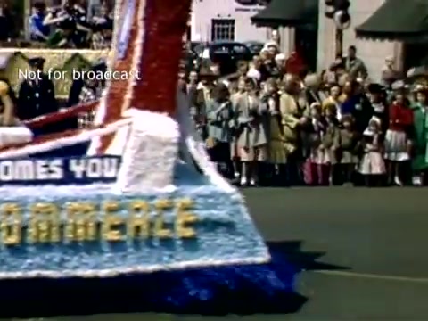 The still depicts a vibrant scene from the late 1940s Holland, Michigan Tulip Festival Parade. A decorated float, prominently featuring the words 'WELCOME YOU,' moves through the streets. Spectators, including families and children in period clothing, line the road, eagerly watching the parade. Colorful decorations and festive elements capture the joyful atmosphere of the celebration.
