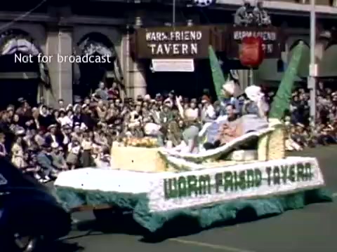 The still captures a vibrant scene from the late 1940s Holland, Michigan Tulip Festival Parade. A colorful float decorates the foreground, featuring floral designs and the name 'Worm Friend Tavern' prominently displayed. In the background, a crowd gathers, eagerly watching the parade, with historic buildings visible behind them. The atmosphere is festive and celebratory, reflecting community spirit and the cultural significance of the event.