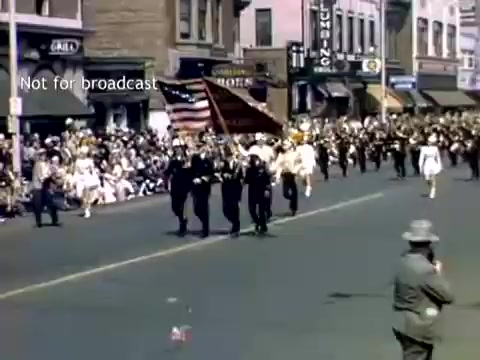 The still captures a lively scene from the late 1940s Holland, Michigan Tulip Festival Parade. A marching group, likely comprised of military personnel, carries an American flag while being greeted by a crowd lining the street. The architecture of the buildings in the background reflects the era, and people are dressed in a mix of festive clothing, indicative of a joyful celebration.