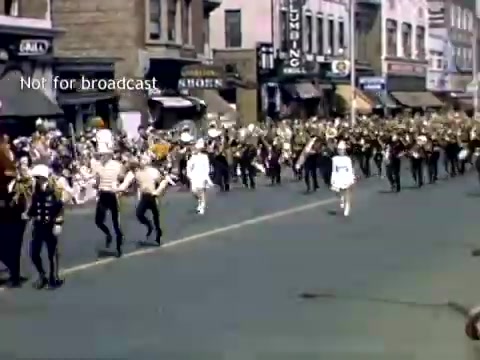 The still captures a lively scene from the late 1940s Holland, Michigan Tulip Festival Parade. A marching band is prominently featured, with musicians dressed in uniforms parading along the street. The festive atmosphere is enhanced by spectators lining the sidewalks, some seated, enjoying the event. Historic buildings can be seen in the background, contributing to the nostalgic charm of the scene.