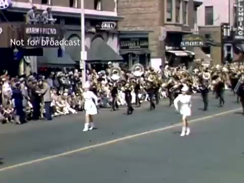 The still captures a lively scene from the Holland, Michigan Tulip Festival Parade in the late 1940s. A marching band is visible, with musicians in uniforms and instruments, while performers in white outfits march in front. A crowded street filled with spectators watching the parade is lined with buildings, evoking a festive atmosphere. The image showcases community celebration and traditional parade festivities.