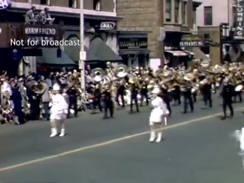 The still captures a lively moment from the Holland, Michigan Tulip Festival Parade in the late 1940s. A marching band, adorned with brass instruments, parades down a busy street, while a crowd watches from the sidewalk. Participants in white costumes add a festive touch to the scene, which is framed by historic storefronts. The atmosphere is cheerful, reflecting the celebratory spirit of the festival.