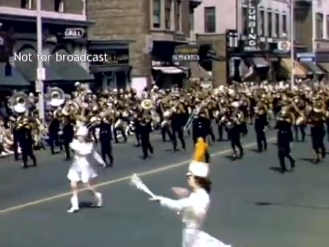 The still captures a lively moment from the Holland, Michigan Tulip Festival Parade in the late 1940s. A marching band, dressed in black uniforms, is prominently featured, performing on a city street. In the foreground, a female majorette in a white outfit leads the parade, twirling a baton. Historic buildings and storefronts line the street, enhancing the festive atmosphere of the event.