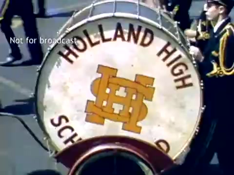 The still features a large drum prominently displaying 'HOLLAND HIGH SCHOOL' in bold letters, with a stylized 'H' logo. The drum is carried by a member of a marching band, likely during the Tulip Festival Parade in Holland, Michigan, in the late 1940s. The scene captures the festive atmosphere of the event.