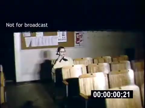 The still appears to show a classroom setting from a 16mm home movie titled 'College Days - 1956.' It features a sparse arrangement of wooden desks, with one individual seated at the front, seemingly engaged in an activity or perhaps on a phone. A bulletin board is visible in the background, adorned with papers and notices. The scene is dimly lit, emphasizing the individual in focus. The text 'Not for broadcast' indicates that this material is not intended for public airing.