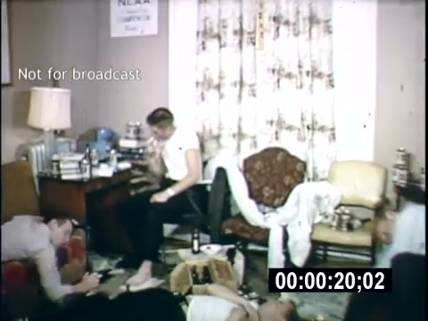 The still shows a casual, cluttered college living room scene from a 1956 home movie. Several young men are present, engaged in relaxed activities. One is sitting at a table with books and drinks around, while others are spread out on the floor and furniture, suggesting an informal gathering. The decor includes a floral curtain and various items scattered around, reflecting a typical college lifestyle of the era. The image has a timestamp overlay indicating it's not for broadcast.