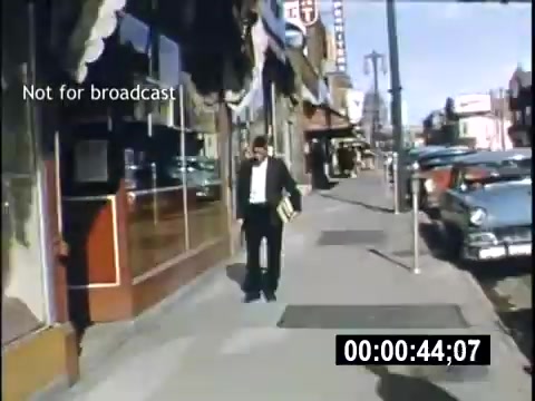The still shows a man walking along a sidewalk, dressed in a suit, with a book under his arm. The scene is set in a college town during the 1950s, with storefronts visible on the left and several vintage cars parked along the street. The bright colors and clear skies suggest a lively atmosphere. The timestamp at the bottom indicates it's from a home movie, capturing a moment of everyday life during that era.