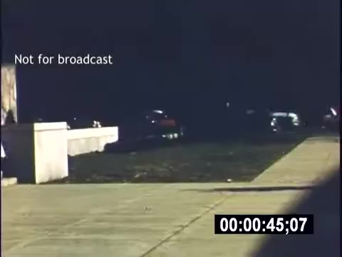 The still shows a college campus scene from a 1956 home movie. There are parked cars visible along a grass area beside a concrete walkway. The image has a vintage quality, with a timecode and a 'Not for broadcast' label. The overall atmosphere captures a moment in student life during the mid-20th century.