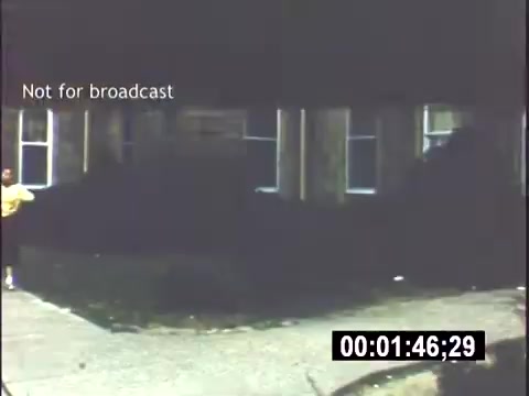 The still captures a scene from a 1956 home movie entitled 'College Days.' It depicts a residential area, possibly near a college campus, with simple buildings and a sidewalk. A person in a yellow shirt stands near the edge of the frame. The lighting is somewhat muted, suggesting it may have been filmed in overcast conditions. The title indicates it is not intended for broadcast.