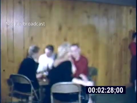 The still captures a casual gathering of young adults in a home setting from the 1956 college days. They are seated around a table, engaged in conversation, with warm wooden paneling in the background. The atmosphere appears relaxed, suggesting a social or friendly gathering.