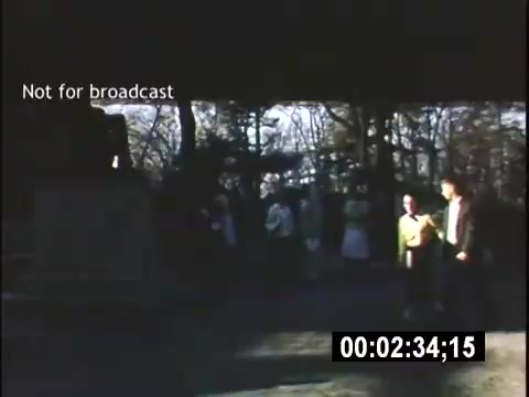 The still from 'College Days - 1956' shows a group of people gathered outdoors, likely on a college campus. The scene is set under a shaded area with trees, suggesting a warm day. A few individuals are interacting, while others are standing slightly apart, possibly engaged in conversation. The image captures a moment of social activity typical of college life in the mid-1950s.