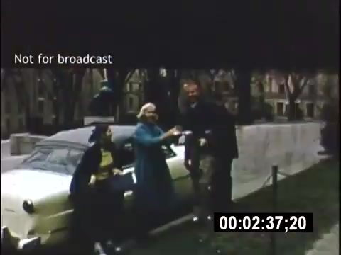 The still captures a scene from the 1956 home movie 'College Days.' It shows three individuals—two women, one wearing a blue coat and the other in a black outfit, and a man—interacting near a parked car. The backdrop features trees and a grassy area, suggesting a college campus atmosphere. The frame is labeled 'Not for broadcast' and includes a timestamp.