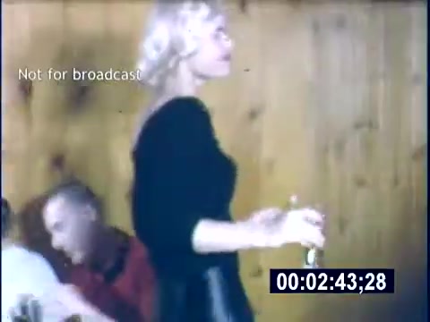 The still shows a woman with blonde hair, wearing a black outfit, walking with a glass in her hand. She is in a casual indoor setting characterized by wooden walls. A couple of people are seated at a table nearby, engaged in conversation. The scene appears lively, likely capturing a social gathering or celebration from the 1956 college era.