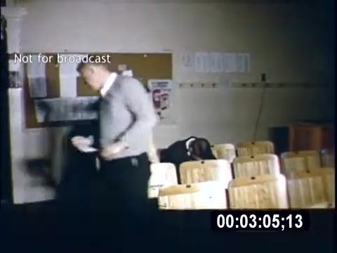 The still captures a scene from the 1956 home movie 'College Days.' In a classroom or lecture hall, a male figure in a sweater is seen walking past rows of empty wooden chairs. Another person, possibly a student, is slouched in a chair, suggesting a relaxed or disengaged atmosphere. The environment appears casual, fitting the theme of college life during that era. A caption indicates the footage is not for broadcast.