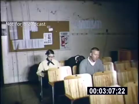 The still features a classroom setting from a 1956 home movie titled 'College Days.' In the image, two students sit at wooden desks, engaged in their studies. The background includes a bulletin board with various notices and posters, while the lighting creates a somewhat subdued atmosphere. The scene captures a moment of academic life in a college environment.