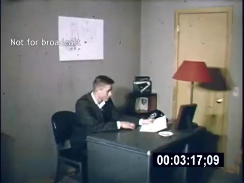 The still shows a young man seated at a desk in a sparsely decorated room. He appears to be engaged with some papers or a typewriter, suggesting a study or administrative setting typical of college life in 1956. The room features a simple lamp, a phone, and a piece of art on the wall. A label indicating 'Not for broadcast' is visible in the corner. The overall aesthetic reflects a mid-century academic atmosphere.