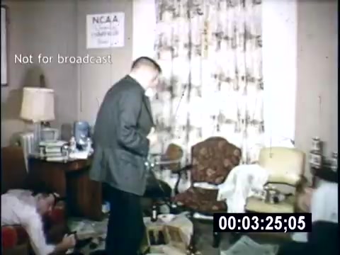 The still appears to show a messy living room scene typical of college life in 1956. A man in a suit stands in the foreground, while another person is partially visible on the floor, suggesting an informal, possibly chaotic atmosphere. The room is cluttered with furniture, books, and papers, capturing a sense of the carefree college experience of that era. A curtain with a floral pattern and a lamp add to the home's cozy yet disorderly feel. The timestamp indicates it is part of a home movie, emphasizing its personal nature.