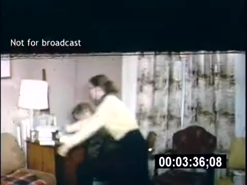 The still from 'College Days - 1956' shows a domestic setting, likely a living room. A woman in a yellow sweater is interacting with a boy, appearing to assist or guide him. There are elements of typical mid-century decor, such as patterned curtains and a chair, contributing to the homey atmosphere. A 'Not for broadcast' label is visible, indicating the footage's private nature. The timestamp suggests it's part of a longer sequence.