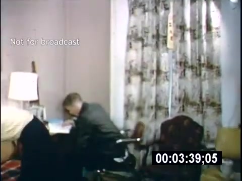 The still shows a room from a 1956 home movie, titled 'College Days.' A young man is seated at a desk, focused on his work, while another figure is partially visible in the foreground, possibly organizing or interacting with the space. The room features patterned curtains and a lamp, suggesting a casual, lived-in atmosphere typical of a college dorm. The image is marked 'Not for broadcast.'