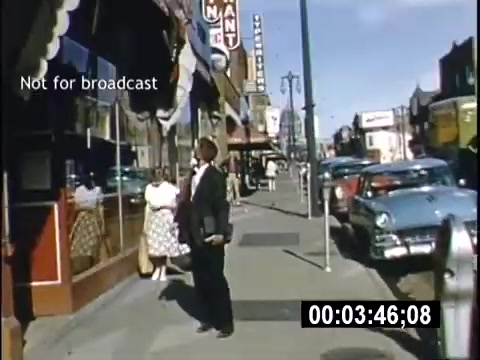 The still from the 1956 home movie 'College Days' shows a sidewalk scene with a man in formal attire gazing into a shop window. Nearby, a woman in a patterned dress stands with her back to the camera. Classic cars are parked along the street, and various storefronts are visible, indicating a vibrant, bustling atmosphere typical of the era. The image captures a nostalgic slice of mid-20th century collegiate life.