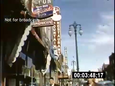 The still from the 'College Days - 1956 home movie' captures a vibrant streetscape featuring storefronts, including a pub and a restaurant, under a clear blue sky. Neon signs are visible, suggesting a lively atmosphere. There are pedestrians along the sidewalk, indicative of a bustling urban environment in the mid-1950s. The scene reflects the everyday life and culture of the time.