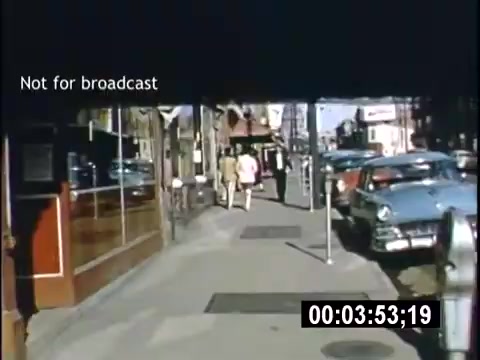 The still features a lively street scene from a 1956 home movie titled 'College Days.' It shows a sidewalk bustling with activity, where two individuals walk together in the foreground. Classic cars line the street, adding to the vintage atmosphere. The scene captures urban life of that era, complete with storefronts and a clear blue sky. A timestamp indicates the moment in the film.