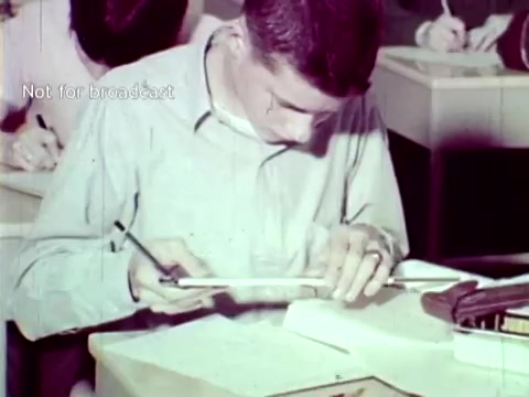 The still from 'IBM - The Information Center (1963)' depicts a student focused on a task at a desk. He is using a tool to measure or draw on a piece of paper, while other students are engaged in similar activities around him. This scene showcases the educational environment where IBM's technologies, like punch cards, are being highlighted for their application in managing academic data such as grades and course enrollment. The retro aesthetic reflects the technological context of the early 1960s.