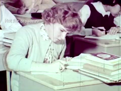 The still from 'IBM - The Information Center (1963)' depicts a classroom scene where a female student is focused intently on her work at a desk, surrounded by stacks of papers. The classroom environment suggests a traditional educational setting, with students engaged in various tasks, highlighting the role of IBM technology in managing academic functions like grades and course enrollment during that era. The image captures a moment of concentrated study, underscoring the intersection of education and emerging computer technology.