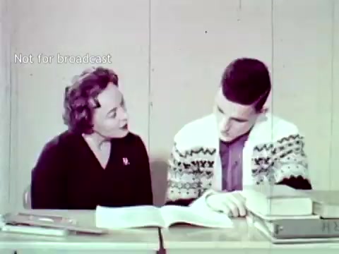 The still from 'IBM - The Information Center (1963)' depicts a woman and a young man seated at a table, engaged in a discussion over a book. The setting suggests an educational context, likely related to course enrollment or grades. The woman appears to be guiding or advising the young man, highlighting the use of IBM technology in academic settings. The vintage aesthetic of the image reflects the era's technological advancements, particularly in computing and information management through methods like punch cards.