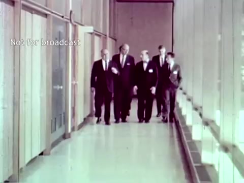 The still from 'IBM - The Information Center (1963)' depicts a group of men walking through a modern corridor, likely within an IBM facility. They are dressed in formal business attire, suggesting a professional setting. The environment is minimalistic, with large windows revealing a bright interior. This scene reflects the cutting-edge technology and corporate atmosphere of the 1960s, emphasizing IBM’s role in advancing educational technology through computers and punch cards for managing grades and course enrollment.