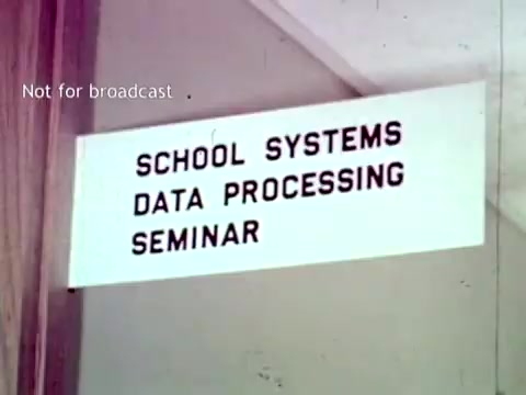 The still features a sign that reads 'SCHOOL SYSTEMS DATA PROCESSING SEMINAR.' It highlights a seminar likely focused on the educational applications of IBM's data processing technology from 1963, indicating a discussion on using such technology for managing school records and administrative tasks. The context suggests an emphasis on the integration of computing into educational administration.