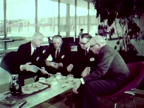 The still from 'IBM - The Information Center (1963)' depicts a group of four men, all dressed in suits, engaged in discussion around a coffee table. They appear to be reviewing documents and possibly using punch cards, highlighting the collaborative nature of IBM's technology in addressing educational administration tasks. The setting, characterized by large windows and modern furnishings, suggests a sophisticated approach to leveraging computer technology for grading and course enrollment.