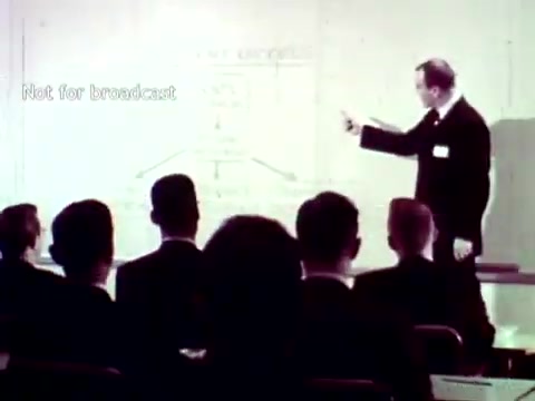 The still captures a scene from the 1963 IBM film, showcasing a speaker, presumably an IBM representative, presenting information to an audience. He points towards a diagram or chart that illustrates the application of IBM technology in educational settings. Attendees, dressed in suits, appear attentive, highlighting the professional context of the presentation. The film emphasizes the innovative use of IBM's punch card systems for managing academic processes like grades and course enrollment. The monochromatic visual style reflects the technological aesthetic of the time.