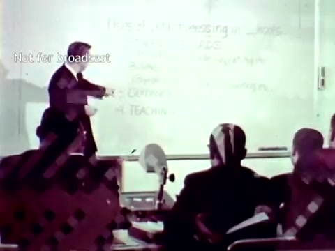The still from 'IBM - The Information Center (1963)' depicts a classroom setting where an instructor is presenting information on a whiteboard. The topic appears to involve data processing related to education, highlighting concepts such as grades and course enrollment. A group of students is seated, attentively watching the presentation, which emphasizes the role of IBM's technology, including methods like punch cards, in modernizing educational processes. The overall aesthetic reflects the early 1960s, showcasing the integration of computer technology into academic environments.