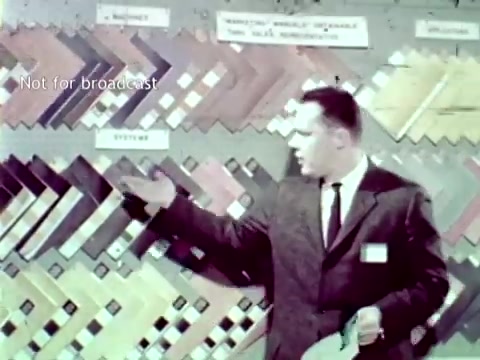 The still from 'IBM - The Information Center (1963)' features a male presenter in a suit, gesturing towards a colorful display of various materials or samples arranged in a geometric pattern on a board. The context suggests that he is showcasing IBM's computer technology and its applications in educational settings, such as managing grades and course enrollment. The overall tone reflects a formal presentation style characteristic of the era.